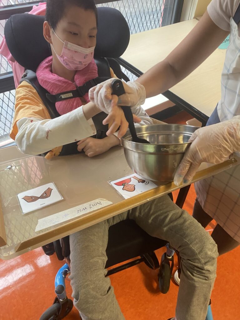 female student in a wheel chair using a spoon to stir and an adult helping her hold the spoon.