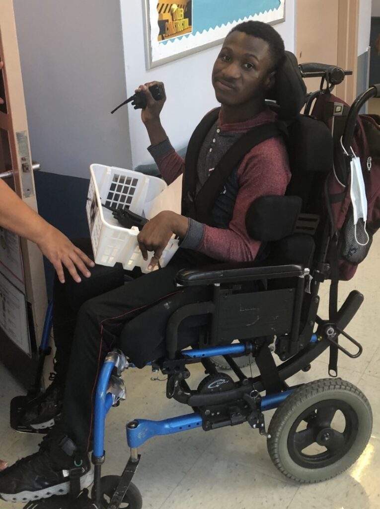 boy in a wheelchair holding a basket in his lap and a walkie talkie in his left hand.