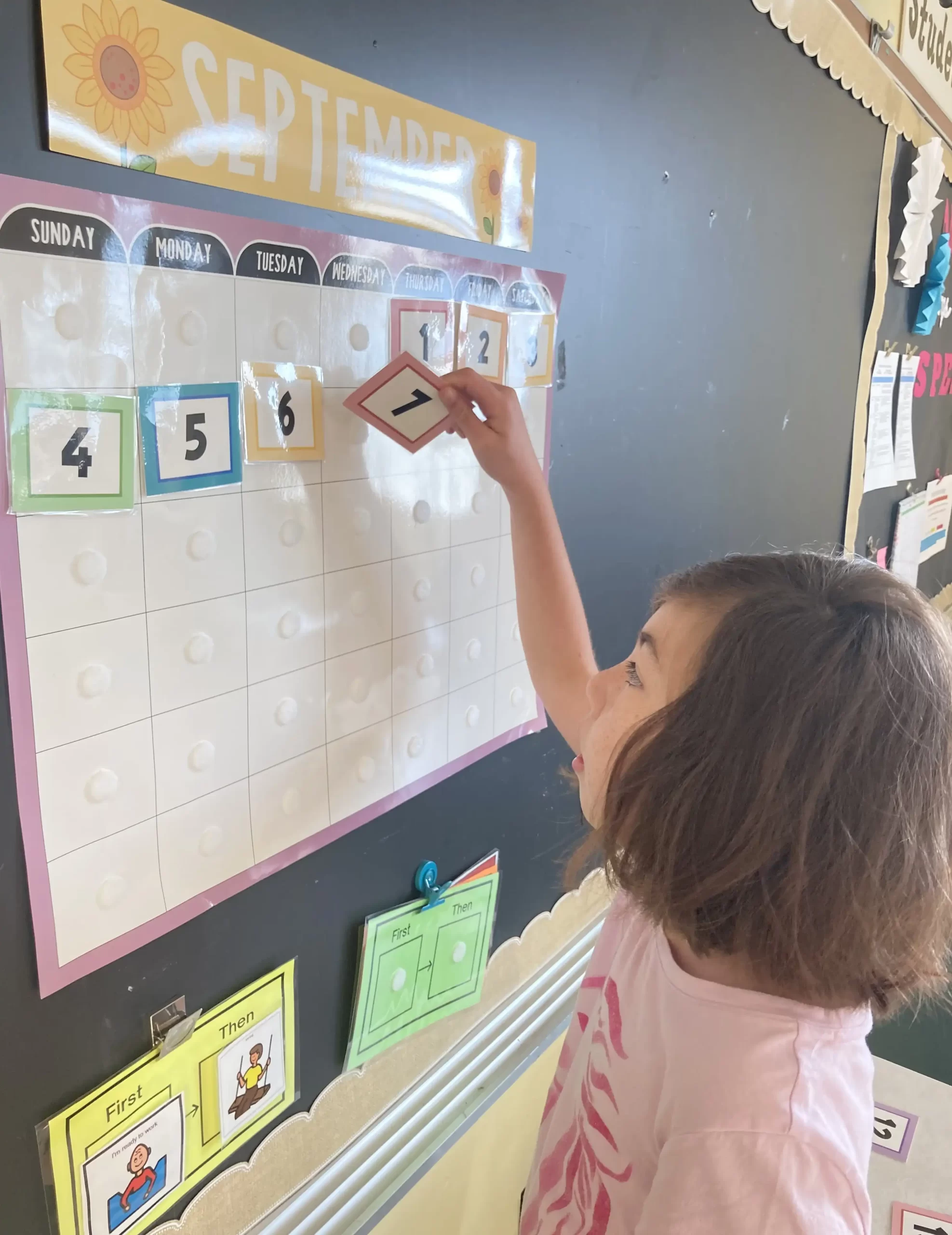 Girl wearing pink shirt placing numbers on a calendar hanging on a blackboard.