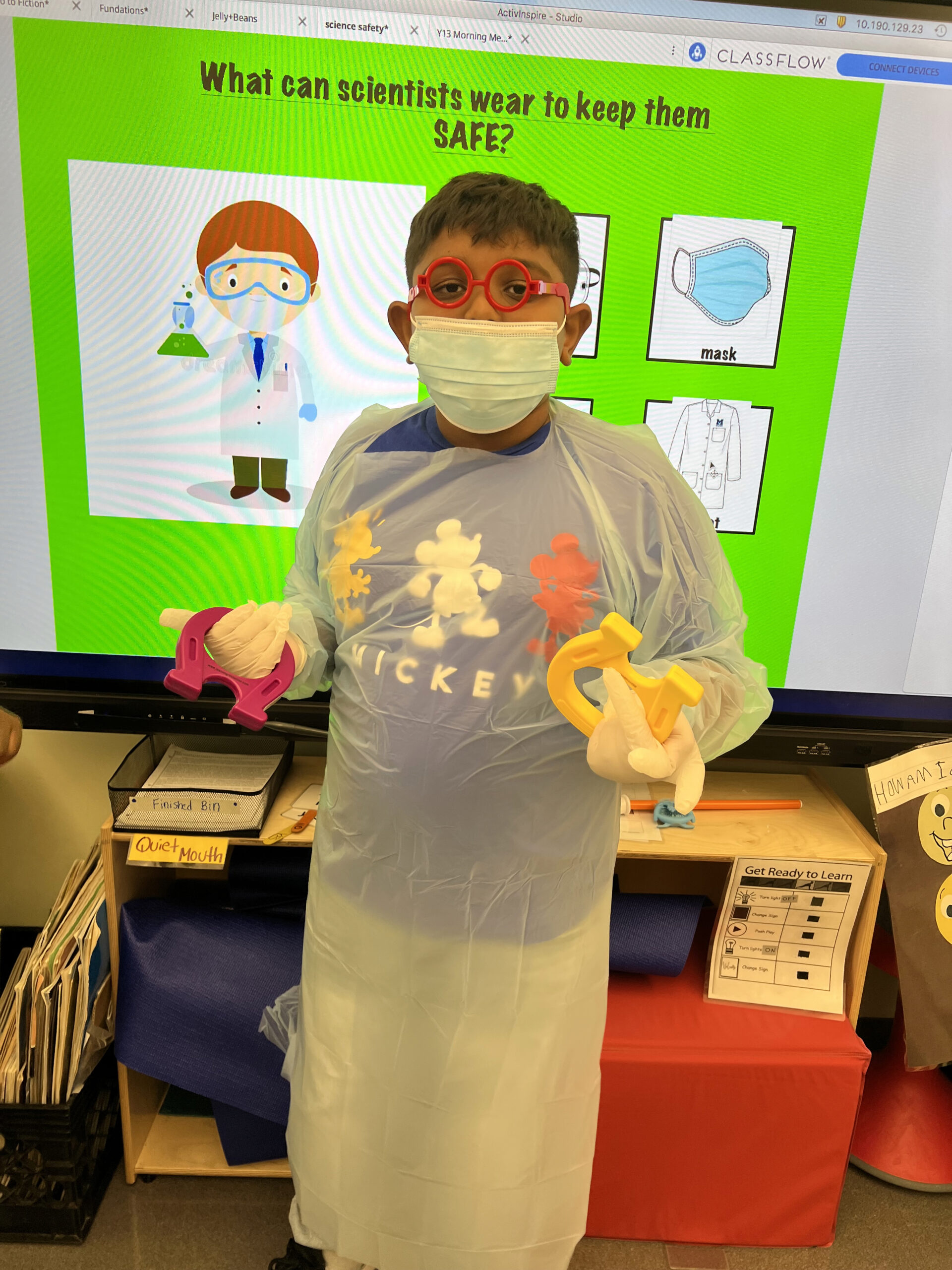 boy wearing lab safety equipment in front o a classroom whiteboard displaying science safety tools goggles, lab coat and gloves