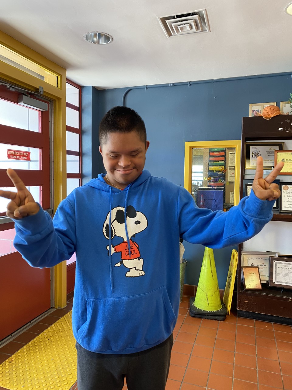 male student standing in his classroom wearing a blue hooded sweatshirt holding two fingers up on each hand.