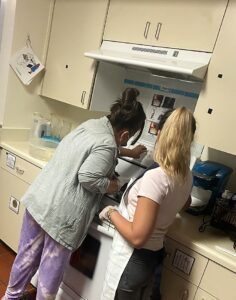 female student and female teacher standing near an oven, the student is turning the oven on.