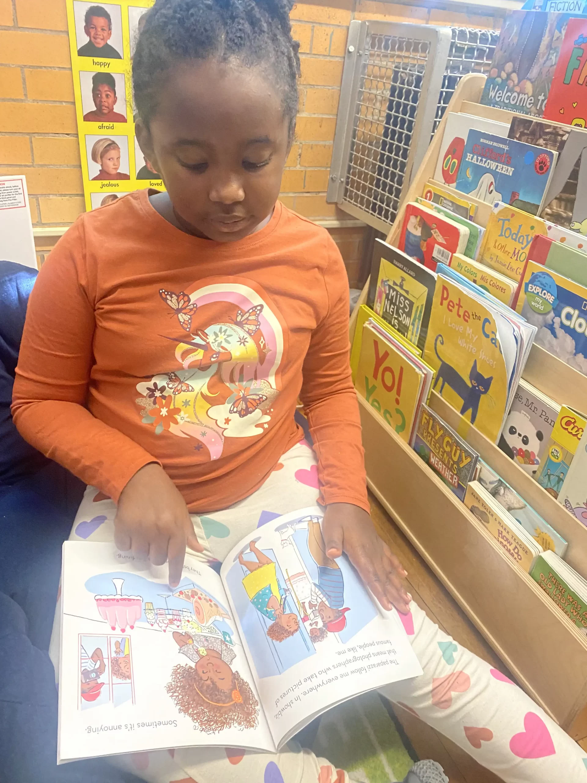 girl wearing an orange shirt reading a book in her classroom.