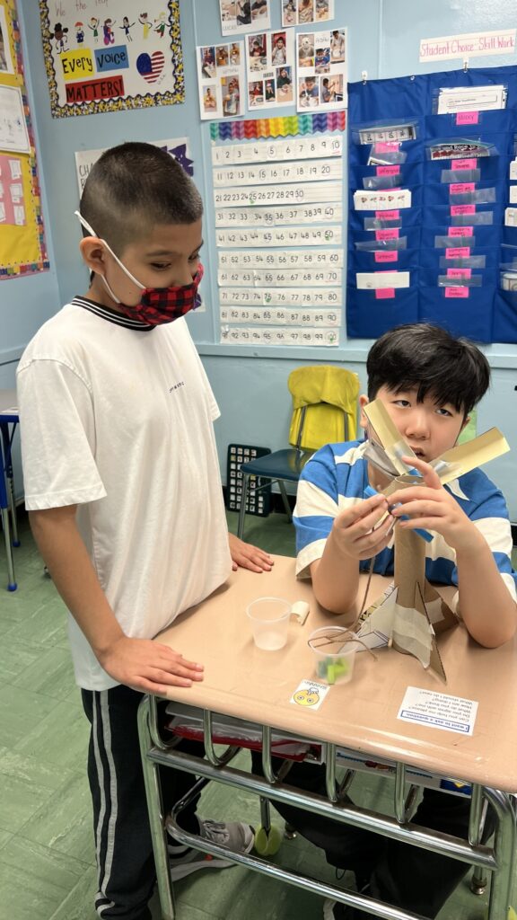 two male students working together to build a windmill made out of a paper towel roll and construction paper.