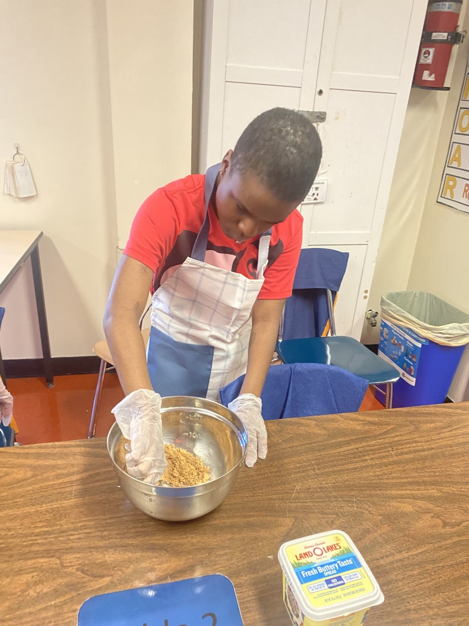 boy wearing a red shirt and white apron using a spoon to stir in a mixing bow.