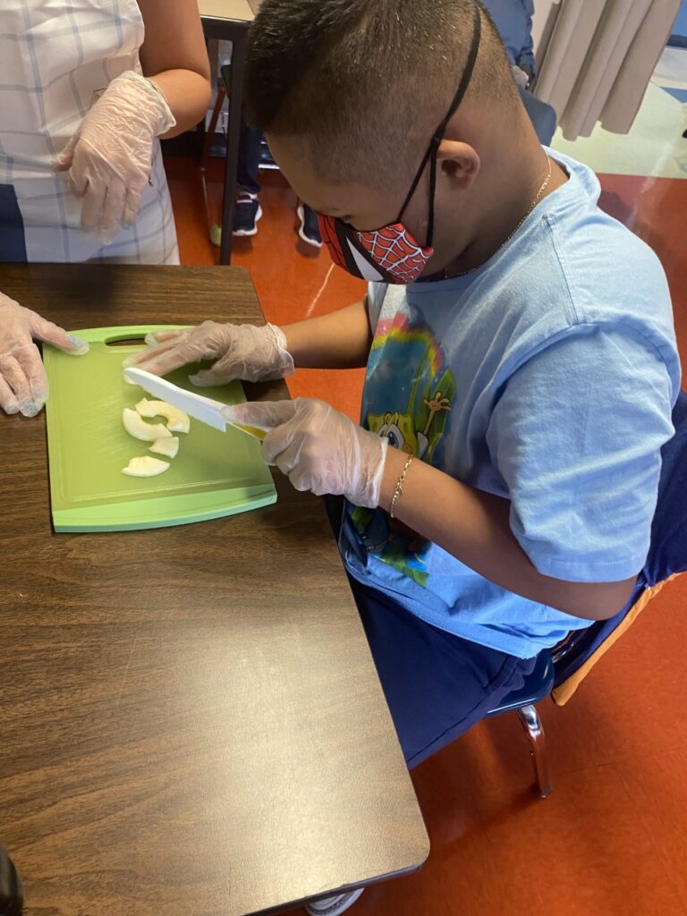 male student with blue shirt and Spiderman face mask cutting apples on a green cutting board.