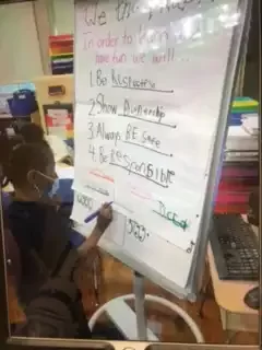 Girl wearing black shirt writing class rules on a large white board.
