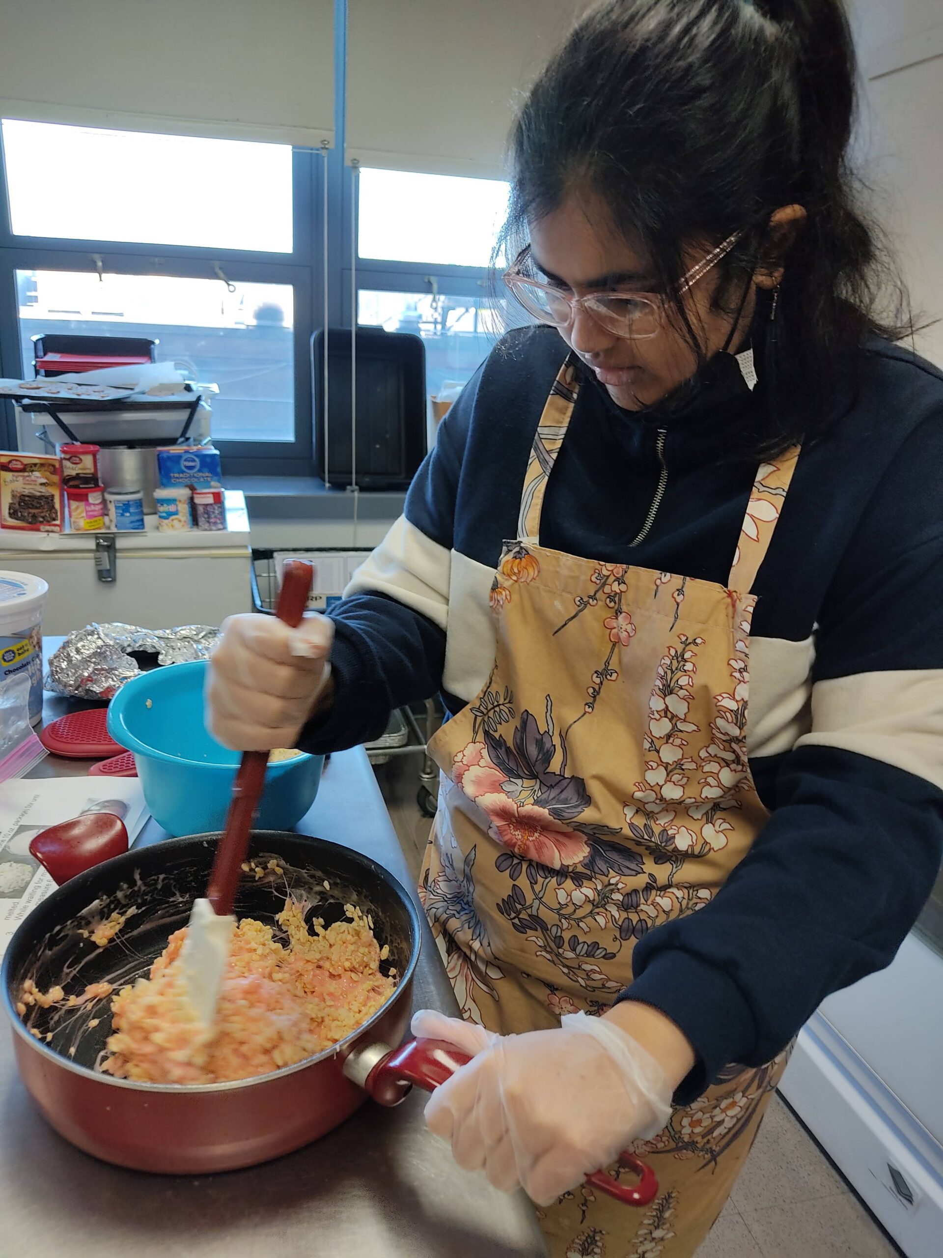 A female student wearing an apron using a spatula to cook food in a frying pan.