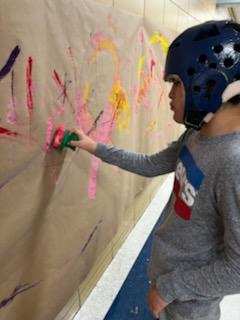 A student wearing a blue helmet using a sponge to paint a poster