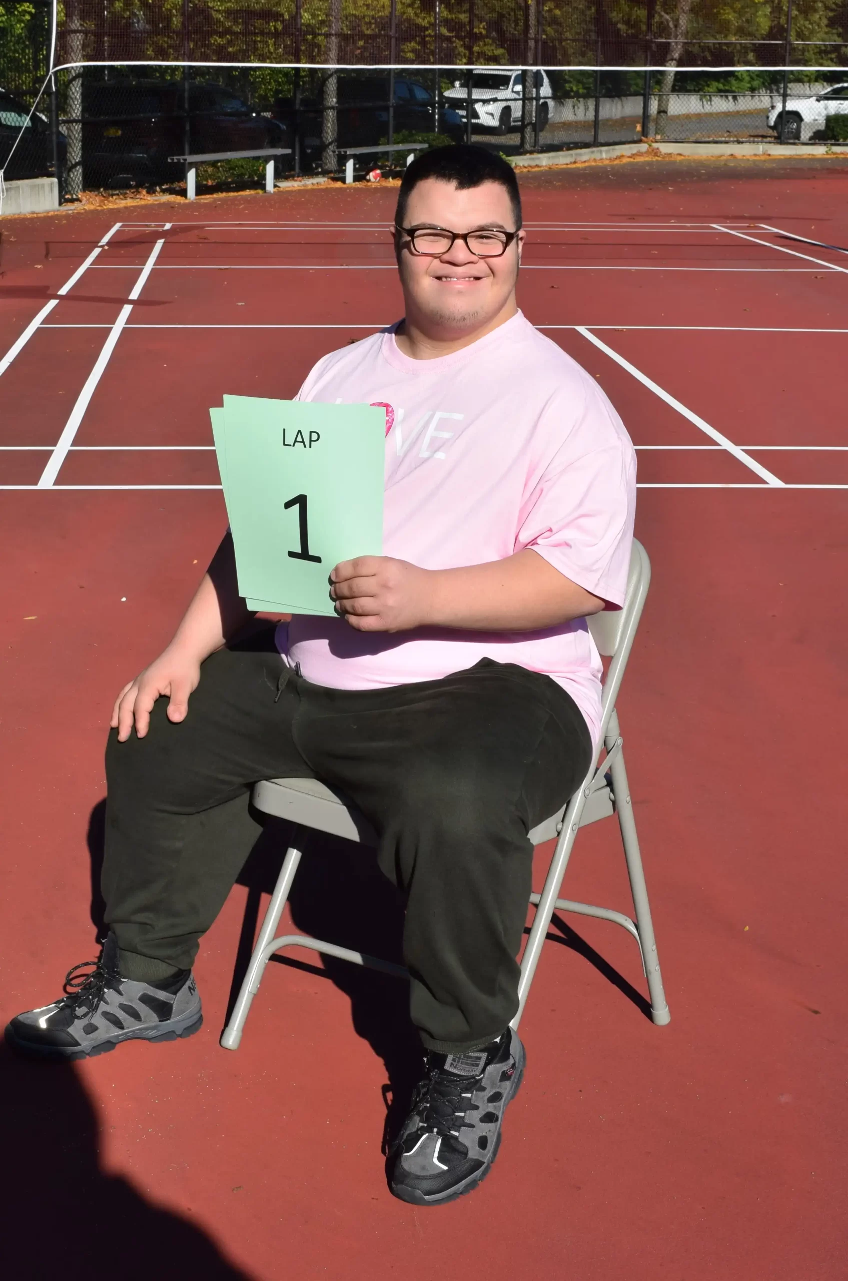 Rayan sitting in a chair wearing a short sleeve pink shirt, helping students keep track of their laps.