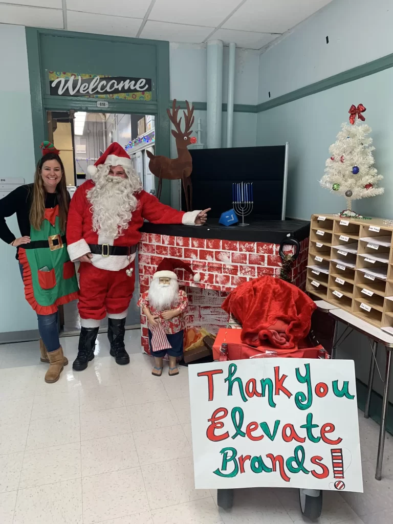 Santa Clause and a female teacher wearing an elf costume standing next to a Menorah.