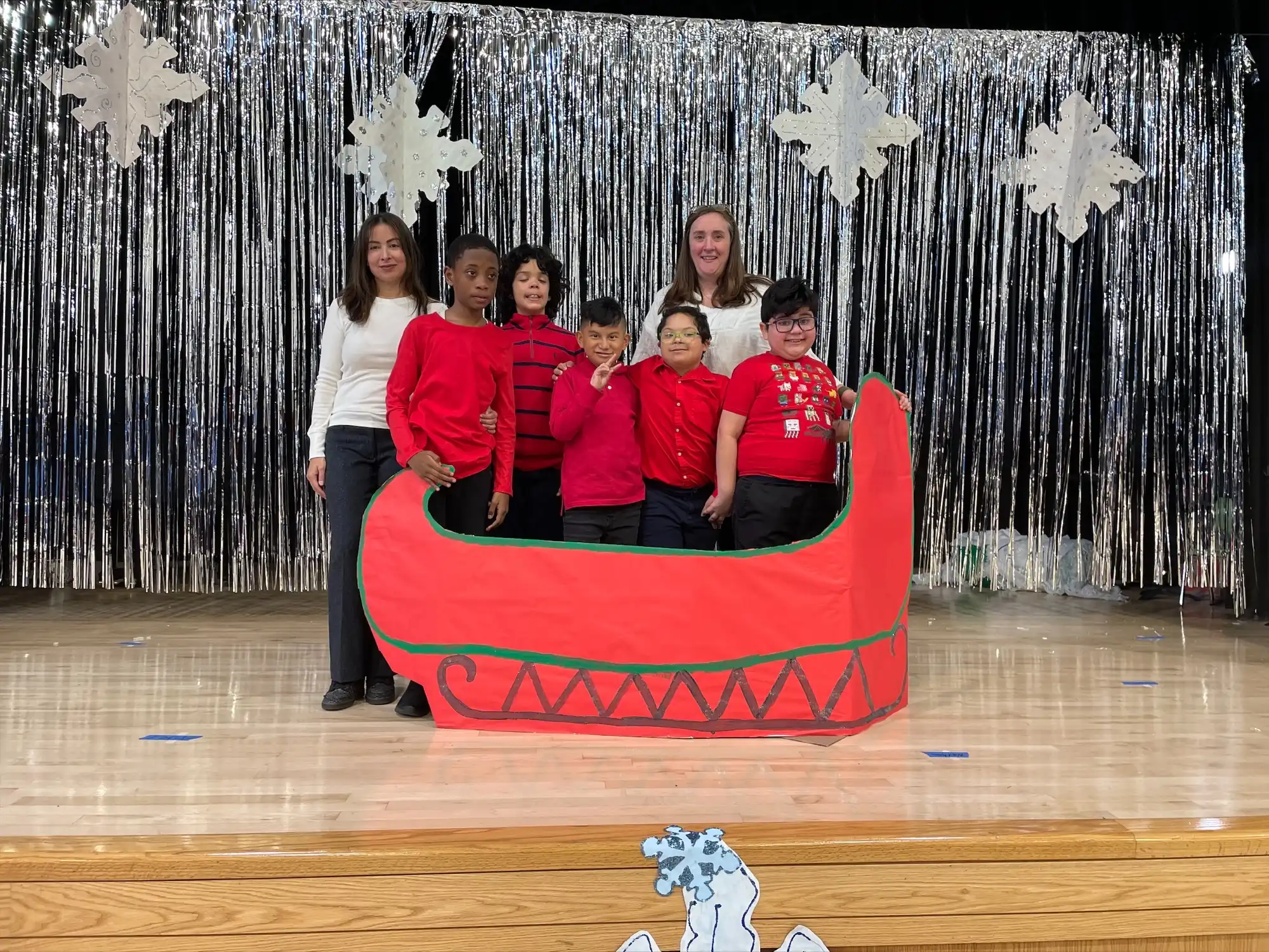 5 elementary school students and two teachers standing next to a cardboard red sleigh.