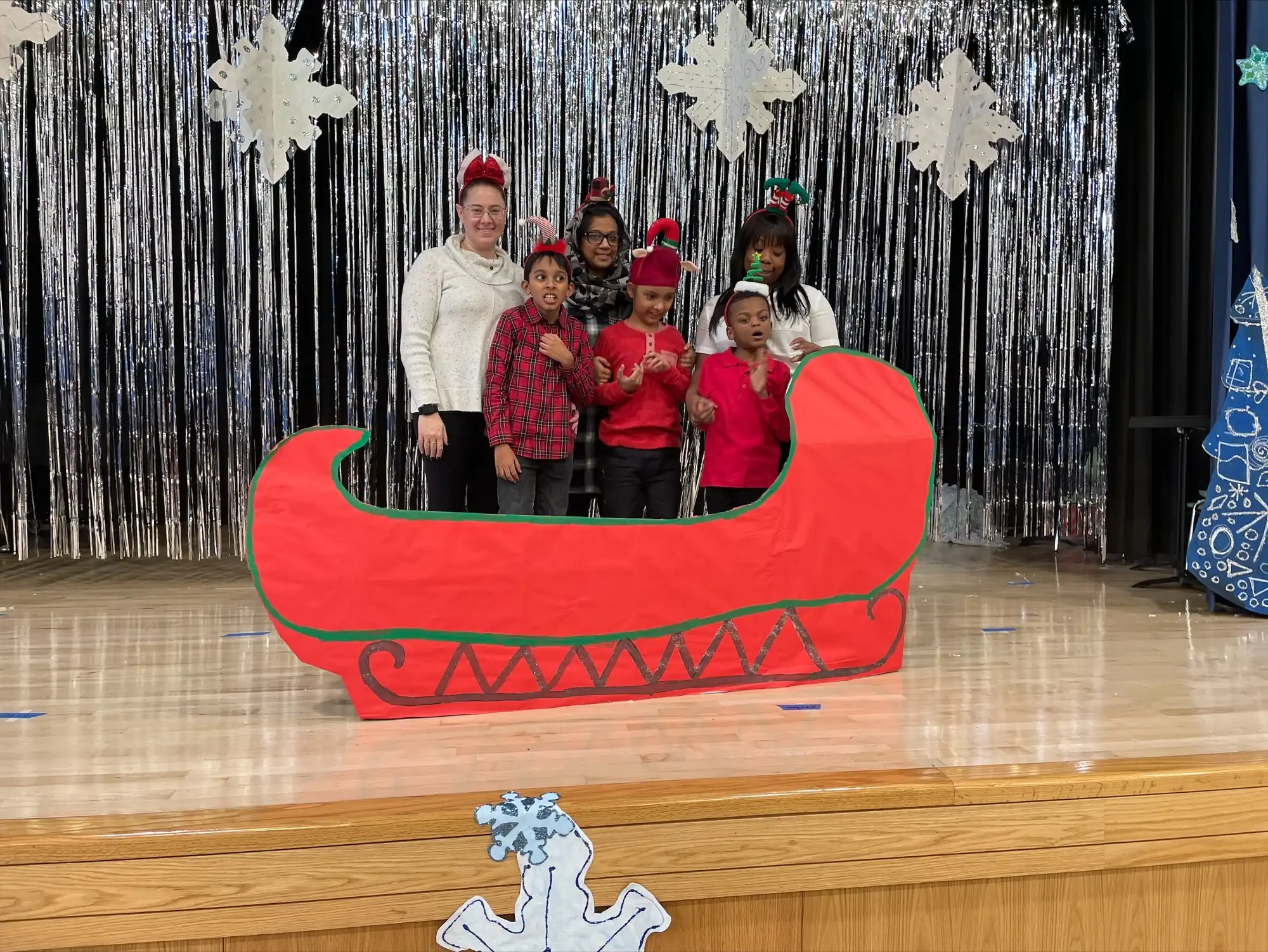3 students and 3 teachers next to a cardboard red sleigh on stage.