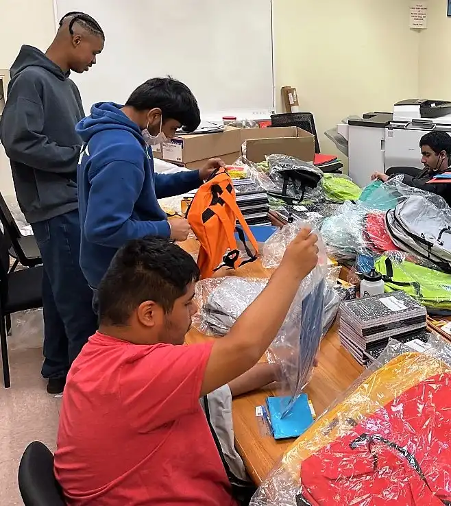 Four ,male students preparing different colored bookbags for distribution during the District 75 bookbag event.