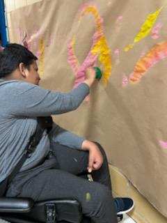 A student in a grey shirt sitting in a wheelchair using a paintbrush to create a pink ribbon