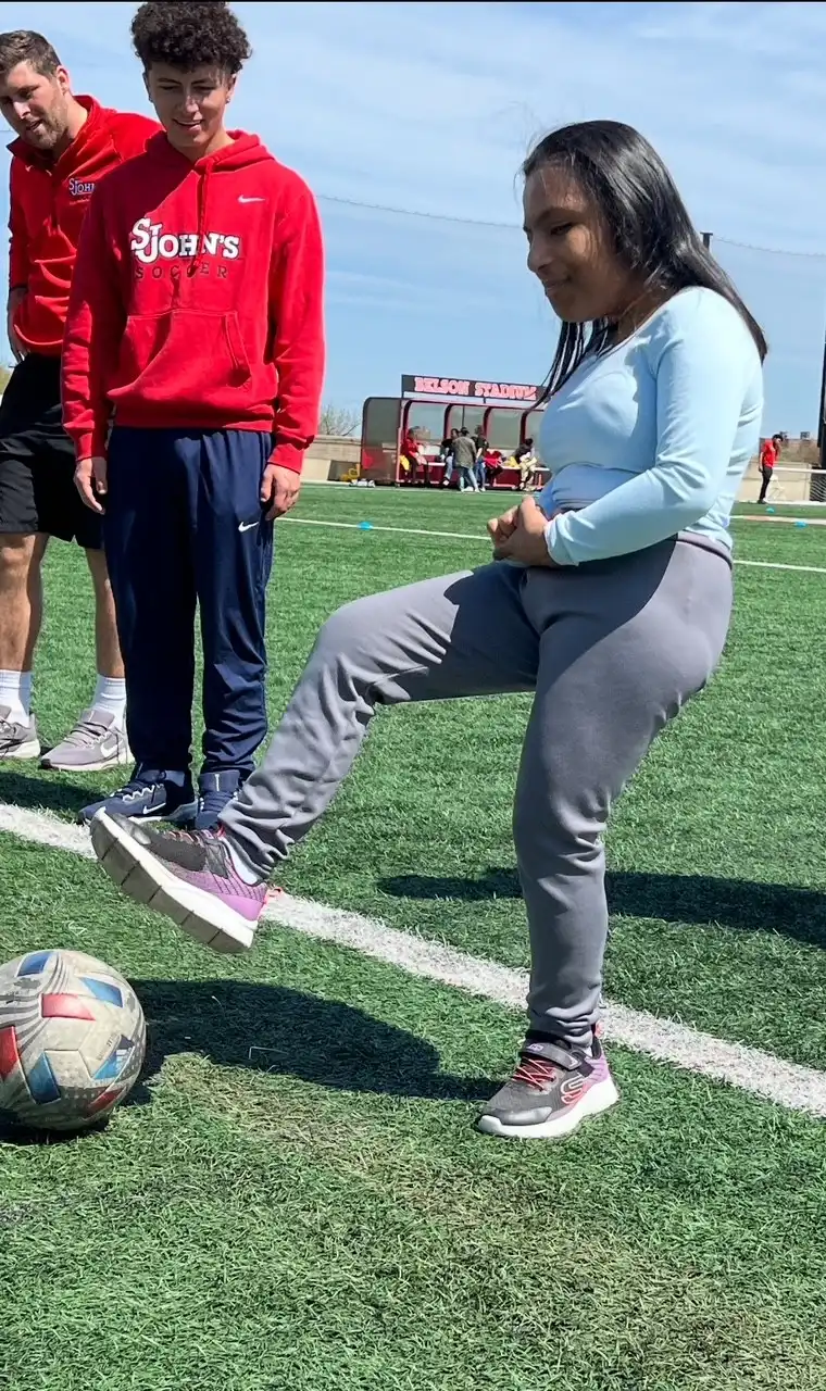 A student wearing a soccer uniform is on a grassy field, mid-kick, sending a soccer ball flying