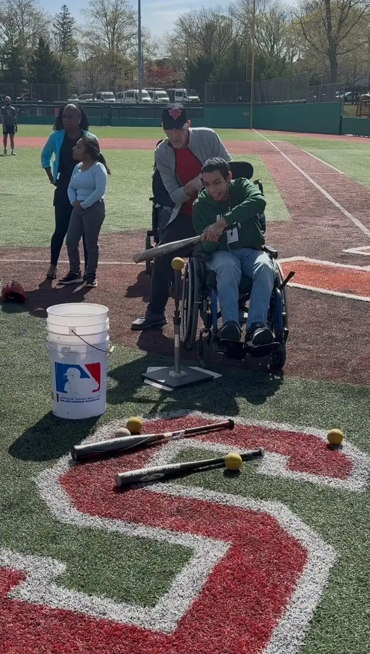 A student in regular clothes is on a grassy field, swinging a bat to hit a ball.