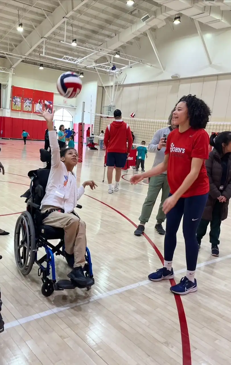 A student in a wheelchair, wearing a green shirt and jeans, is in a gym throwing a ball to a St. John's player.