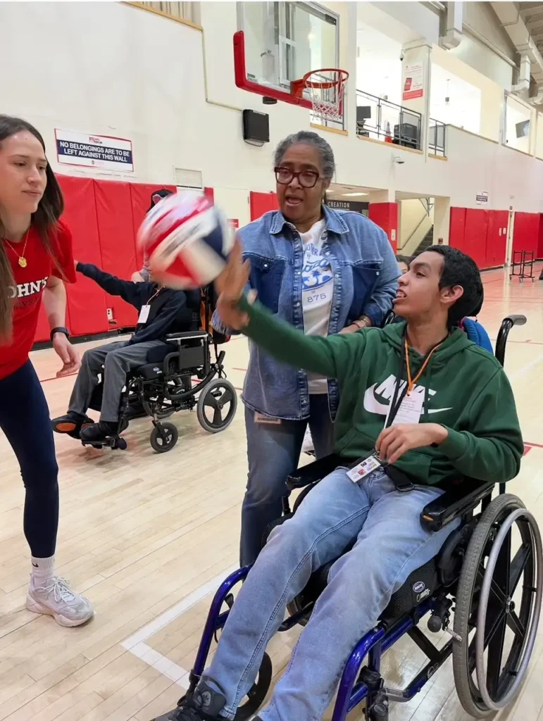 "A student in a green shirt and jeans is in a wheelchair on a basketball court, throwing a basketball to another player."