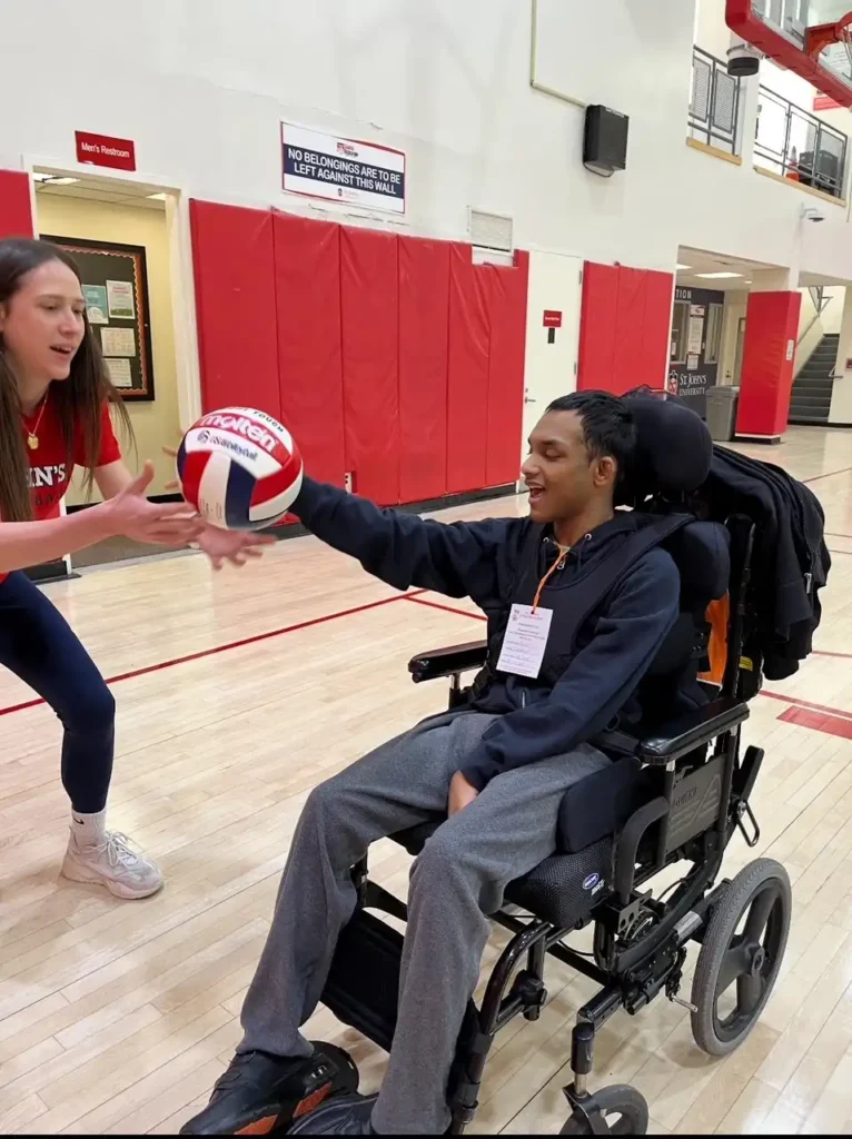 "A student in a blue shirt is in a wheelchair on a basketball court, throwing a basketball to another player."