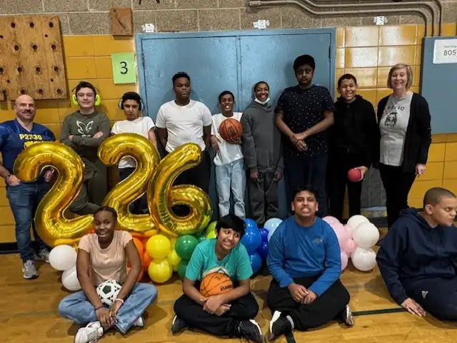 “A group of students and teachers stand together smiling in front of large yellow balloon numbers that read ‘226,’ with colorful balloons arranged beneath the numbers.”