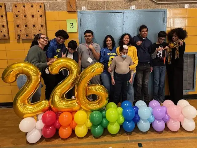 “A group of students and teachers stand together smiling in front of large yellow balloon numbers that read ‘226,’ with colorful balloons arranged beneath the numbers.”