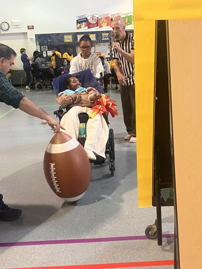A student in a manual wheelchair leans forward to kick a brightly colored, oversized inflatable football on a grass field during a school gym activity.