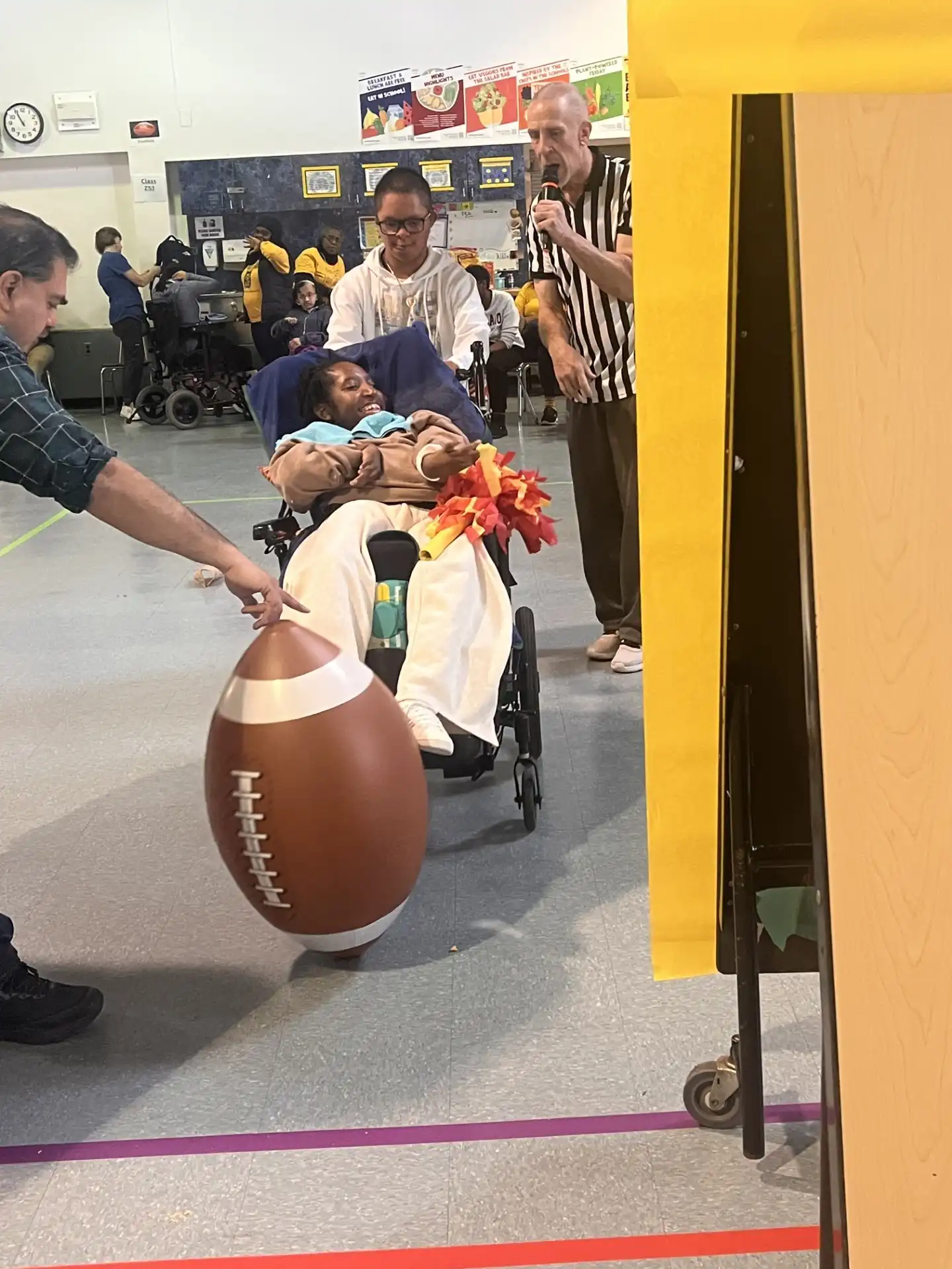 A student in a manual wheelchair leans forward to kick a brightly colored, oversized inflatable football on a grass field during a school gym activity.