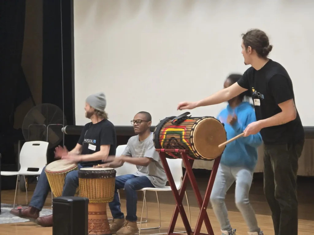 A student participating in a live cultural performance, playing a djembe drum on stage with a West African drum ensemble during a school assembly.