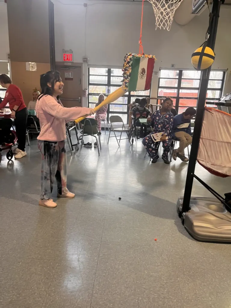"Students enjoying a festive cultural activity in the gym, featuring a student attempting to break a piñata hanging from a basketball hoop.