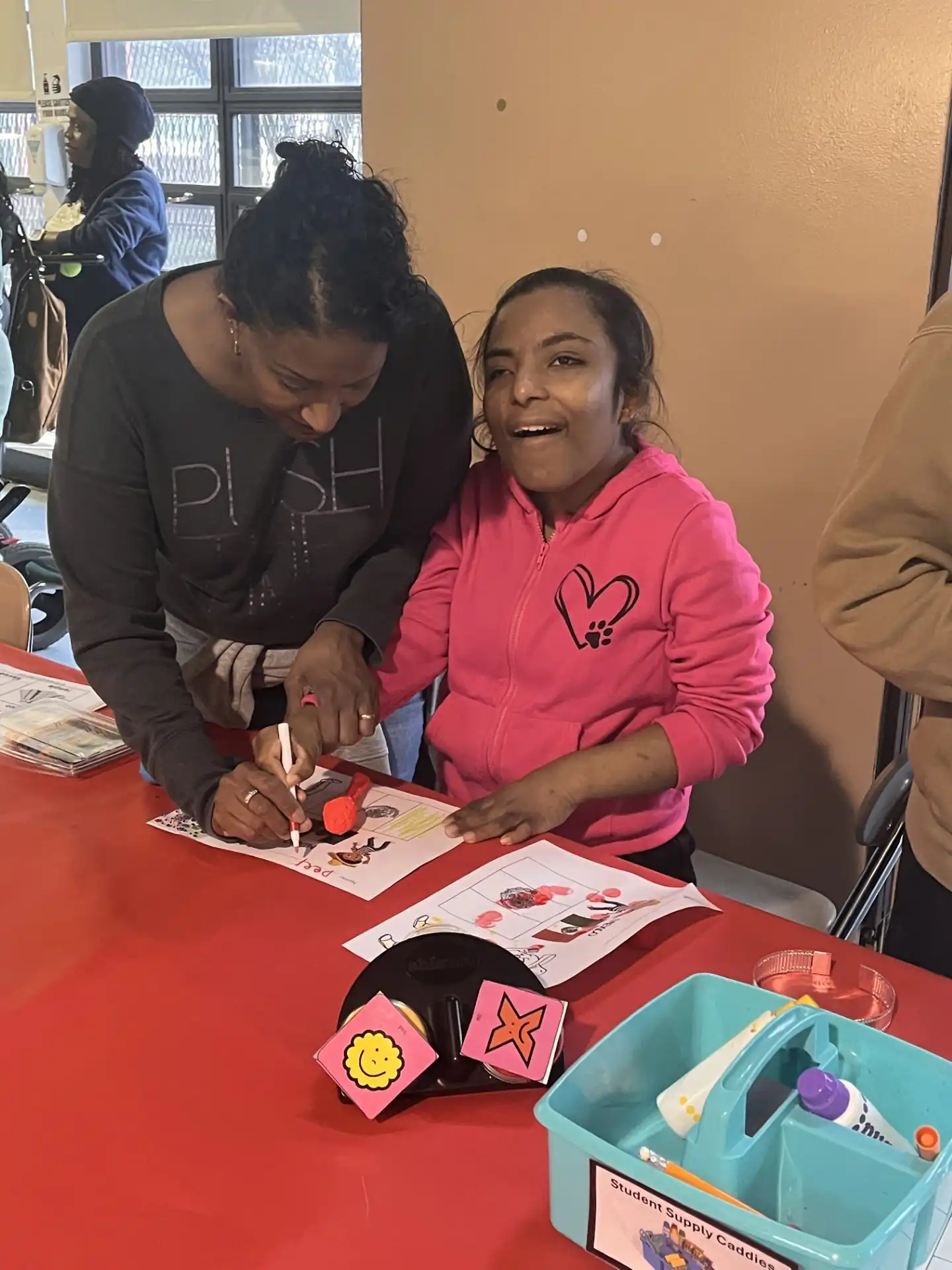 "A teacher leans over a student's desk to provide guidance as the student writes on a printed worksheet. The teacher is pointing to a specific section of the page while the student looks on intently in a classroom setting.