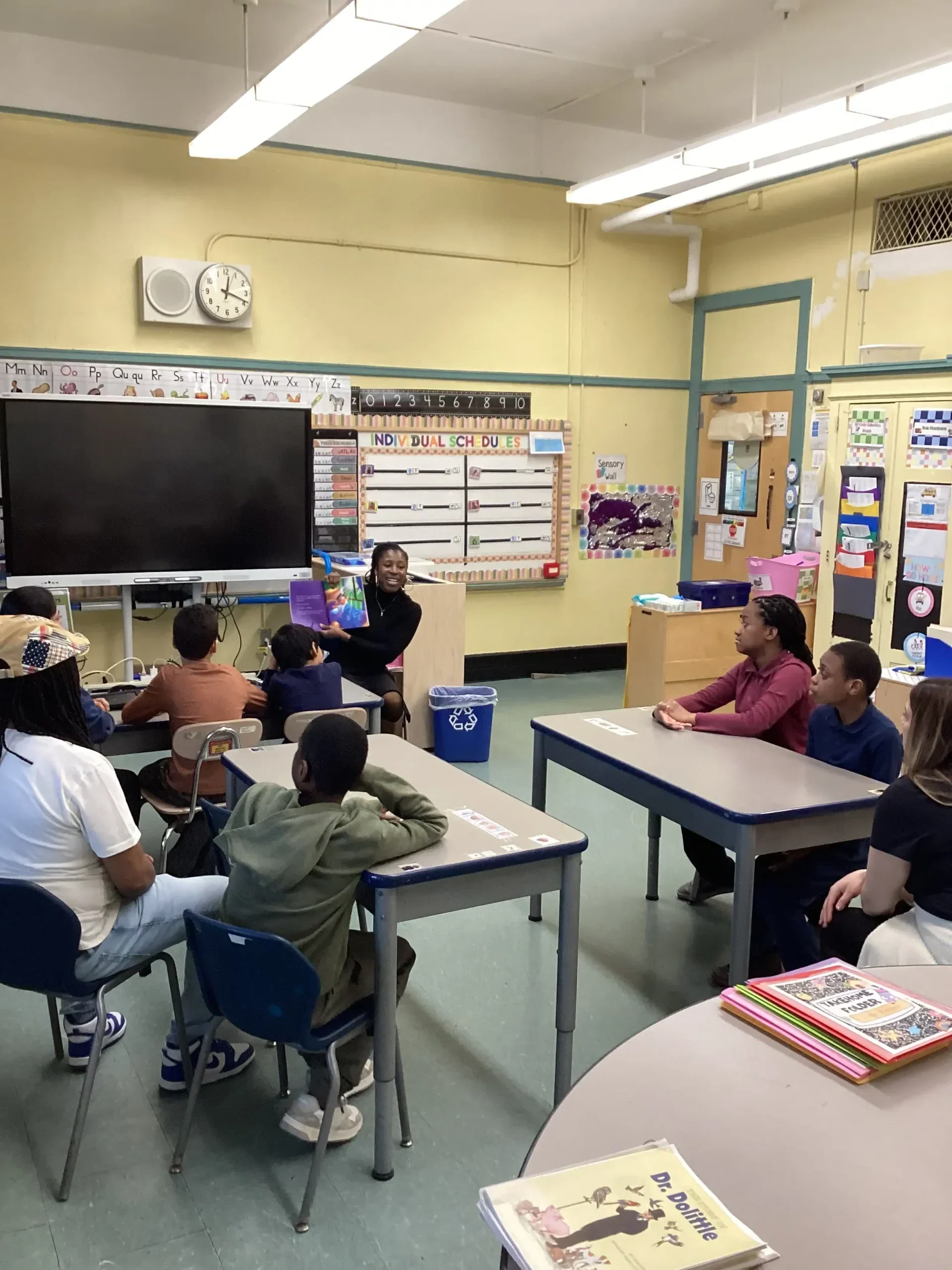 A student stands at the front of a classroom, holding a book and reading aloud to their classmates who are seated at desks and listening attentively