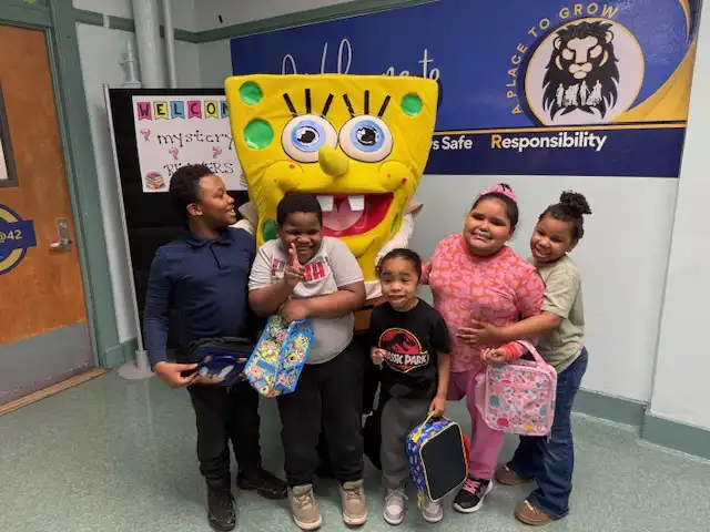 Mascot character SpongeBob SquarePants reading a story to a captivated group of elementary school students during a literacy event.