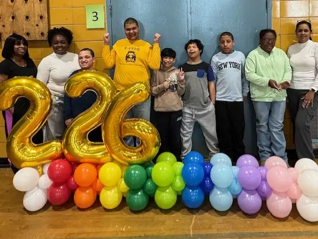 “A group of students and teachers stand together smiling in front of large yellow balloon numbers that read ‘226,’ with colorful balloons arranged beneath the numbers.”