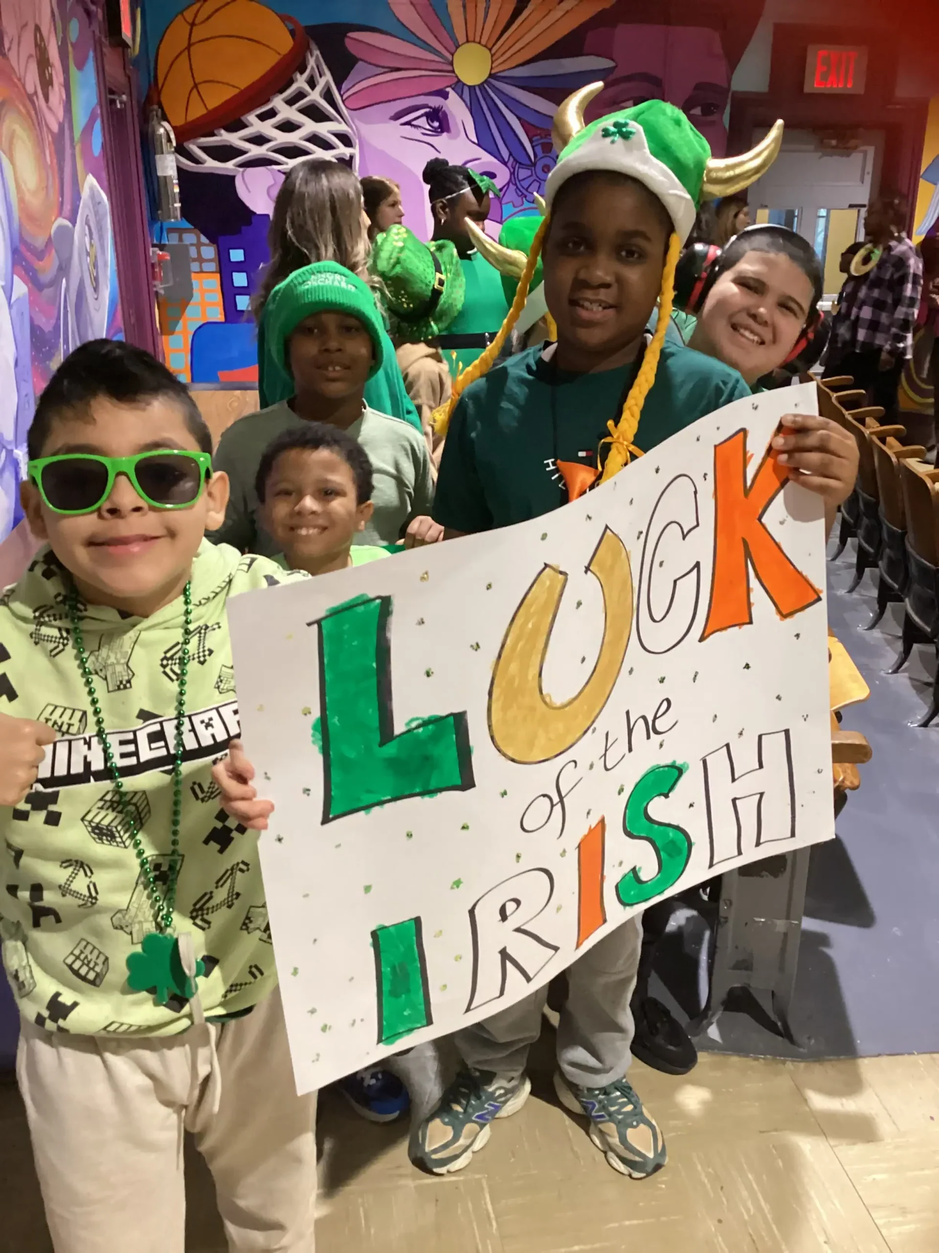 Five students dressed in green and festive St. Patrick’s Day colors, smiling and holding a holiday-themed sign.